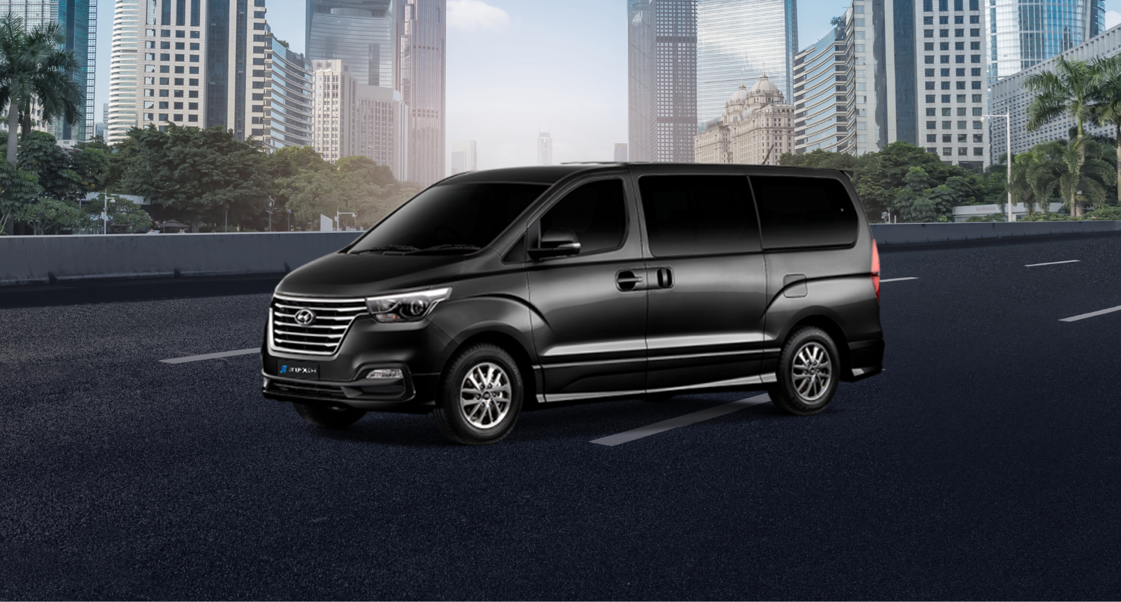 A sleek, black Hyundai H1 van is parked on an asphalt road, set against a backdrop of urban skyscrapers and trees. The van features tinted windows for privacy and silver alloy wheels.