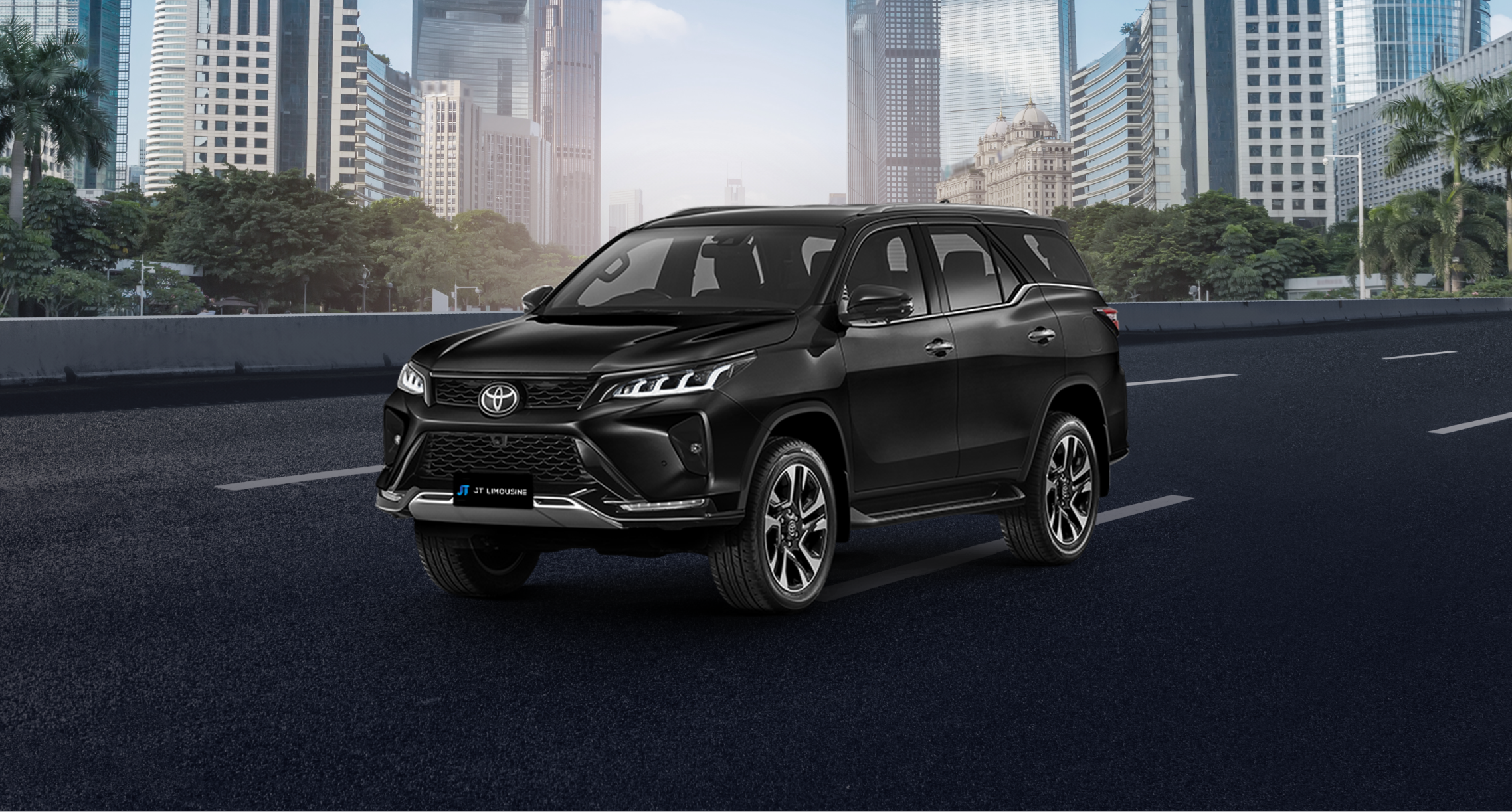 A sleek, black Toyota Fortuner SUV is parked on an asphalt road, set against a backdrop of urban skyscrapers and trees. The vehicle features slim LED headlights, a large black grille, and two-tone alloy wheels. 