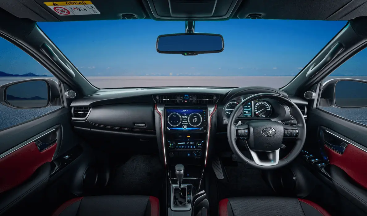 An interior view of a current-model Toyota Fortuner or similar vehicle, looking from the back seats towards the front. The cabin features a dark color scheme with deep red accents on the door panels and parts of the seats. A large central touchscreen display shows navigation and infotainment, while a multi-function steering wheel, a digital/analog instrument cluster, and a rearview mirror are visible. Outside the windshield, a vast desert or open landscape stretches under a clear blue sky.