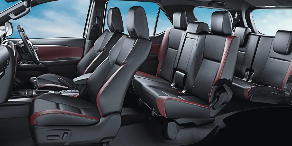 An interior view of a Toyota Fortuner, highlighting the full three rows of passenger seating. The seats are black with deep red accents or borders and feature red stitching details. The door panels also maintain the black and red color scheme consistent with the seats, showcasing a sporty and luxurious interior design.