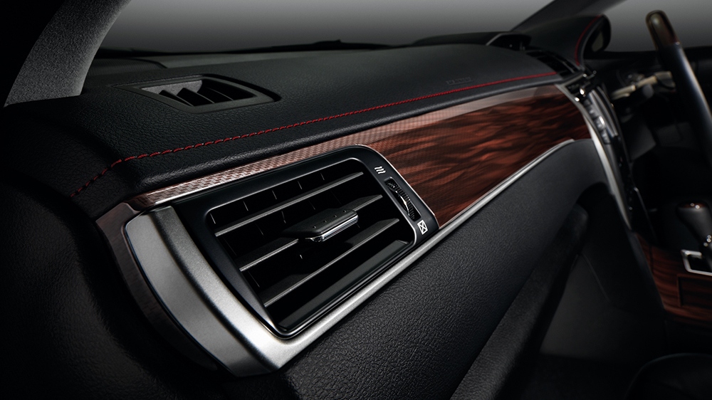 An interior shot of a Toyota Camry, focusing on the passenger side dashboard. It features prominent rectangular air vents with chrome trim, accented by a reddish-brown wood grain panel and red stitching on the black console.