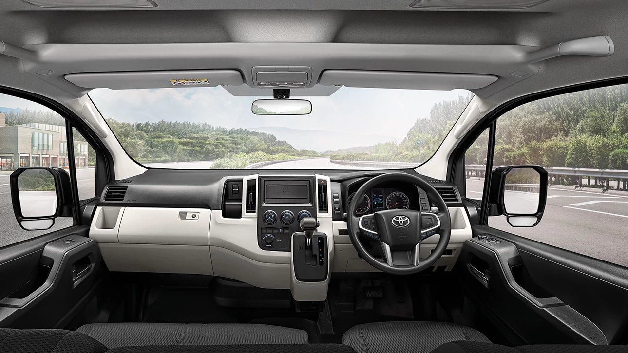 A wide-angle shot of the front interior of a white and black Toyota Commuter van, featuring a right-hand drive steering wheel. The dashboard includes an infotainment screen centered above two small air vents. The driver's instrument cluster shows a speedometer and tachometer. Side mirrors are visible through the left and right windows, with an outside view of a road lined with trees and buildings.