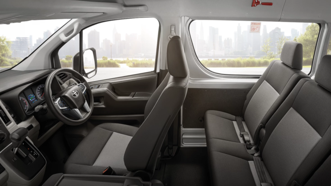 A wide-angle shot of the interior of a Toyota Commuter van, showing both the passenger and driver's seating area. There are three rows of passenger seats upholstered in dark and light grey fabric. The first row of passenger seats features adjustable headrests, and the second and third rows are horizontally foldable. The van's steering wheel is on the right side, indicating a right-hand drive vehicle. The van's windows reveal a distant city skyline. All rows of seats are equipped with seatbelts for passenger safety.
            