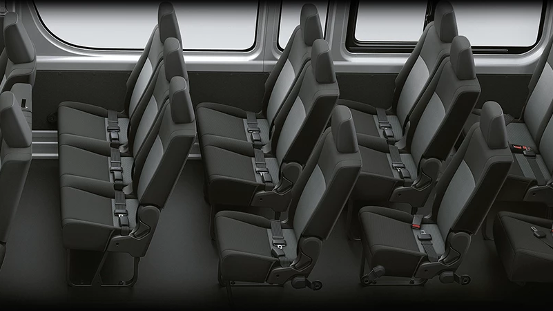 An elevated view of the rear cabin of a passenger van, showing multiple rows of passenger seats facing the left side of the image. All seats are upholstered in black and grey fabric and have seatbelts. Light streams in from large windows above the seats, revealing a slightly blurred view of buildings outside. There is an aisle or space for luggage on the left side of the image.