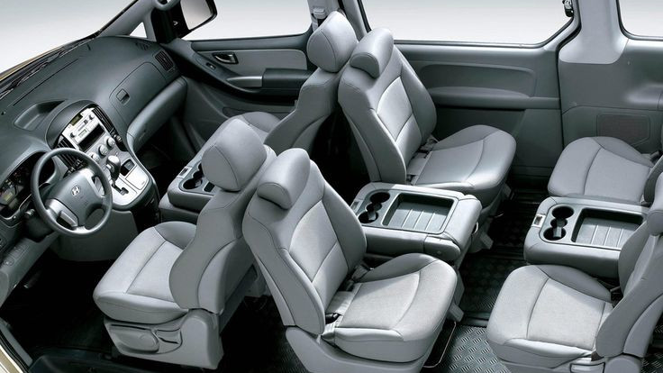 An interior view of a multi-purpose vehicle (MPV) or van, looking from the rear towards the front, showcasing numerous light gray passenger seats. The seating arrangement appears flexible, with some seats folded and integrated cup holders. The floor of the cabin features a textured, possibly carpeted or rubberized, surface for durability.