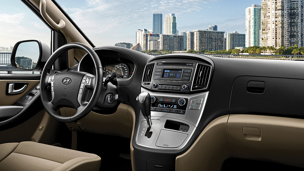 An interior view of a Hyundai H1 or Hyundai H1 Staria from the driver's perspective, showing a two-tone beige and black dashboard, a central infotainment and display screen, rotary climate control knobs, and a multi-function steering wheel. Outside the windshield, a cityscape with high-rise buildings is visible.