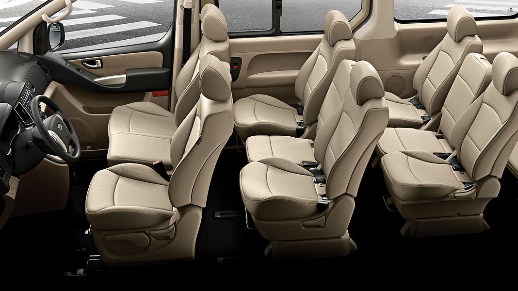 An interior view of a Hyundai H1 or Hyundai H1 Staria, showcasing multiple rows of passenger seating within the cabin. All seats are beige or cream-colored, contrasting with the dark floor. The interior appears spacious and designed to accommodate a large number of passengers.